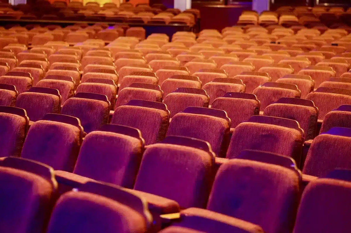 Escena de teatro al aire libre con público sentado y escenario iluminado al atardecer